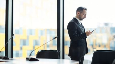 In Politik und Wirtschaft finden Kryptohandys Verwendung Ein Mann im Anzug steht in einem Raum mit einem Tisch vor einer Fensterfront und blickt auf sein Mobiltelefon