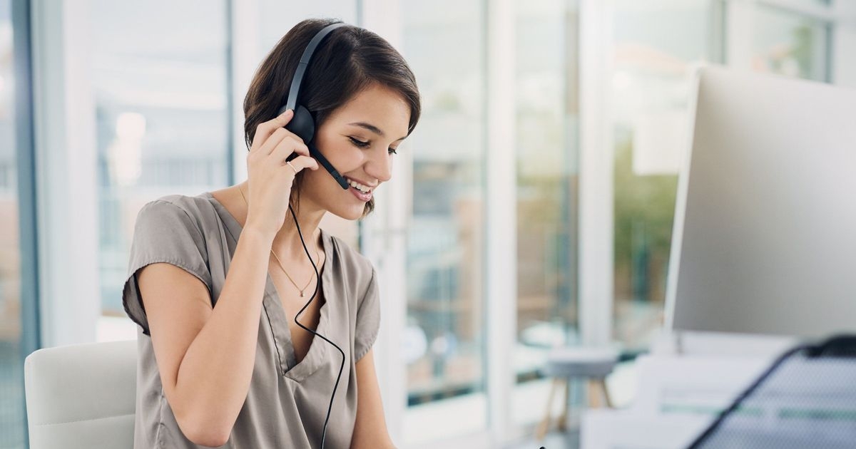 Eine Frau sitzt an einem Tisch und trägt ein Headset. Mit der rechten Hand fasst sie an den Kopfhörer, in der linken Hand schreibt sie mit einem Stift auf einen Notizblock.