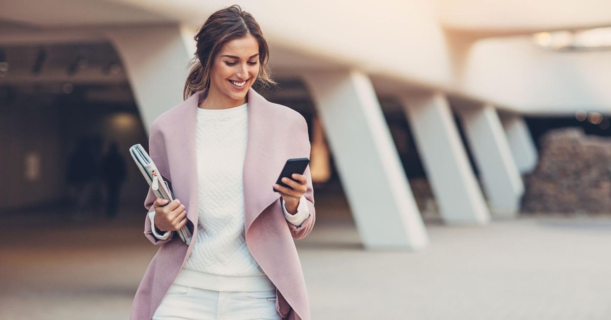 Eine Frau mit einer rosa Jacke steht im Freien vor einem Gebäude und betrachtet lächelnd ihr Mobiltelefon
